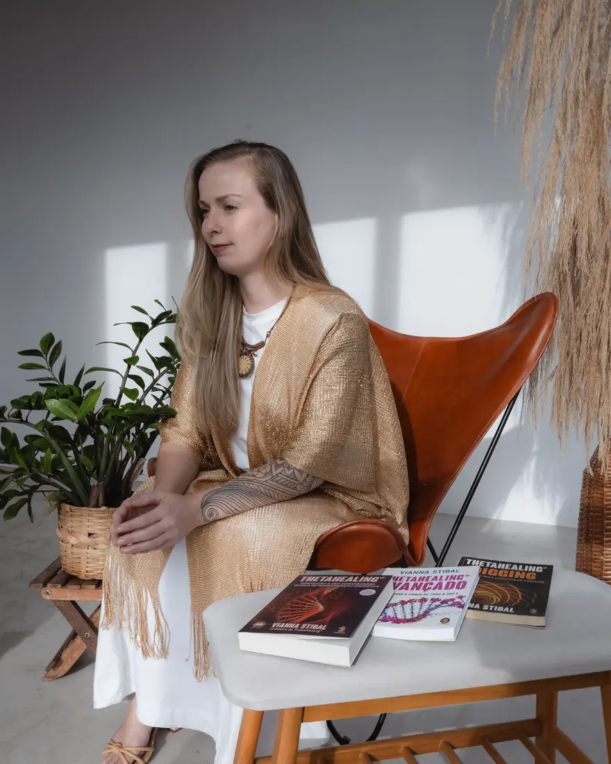 Lairany Souza sentada em uma poltrona ao lado de uma mesa com livros de ThetaHealing