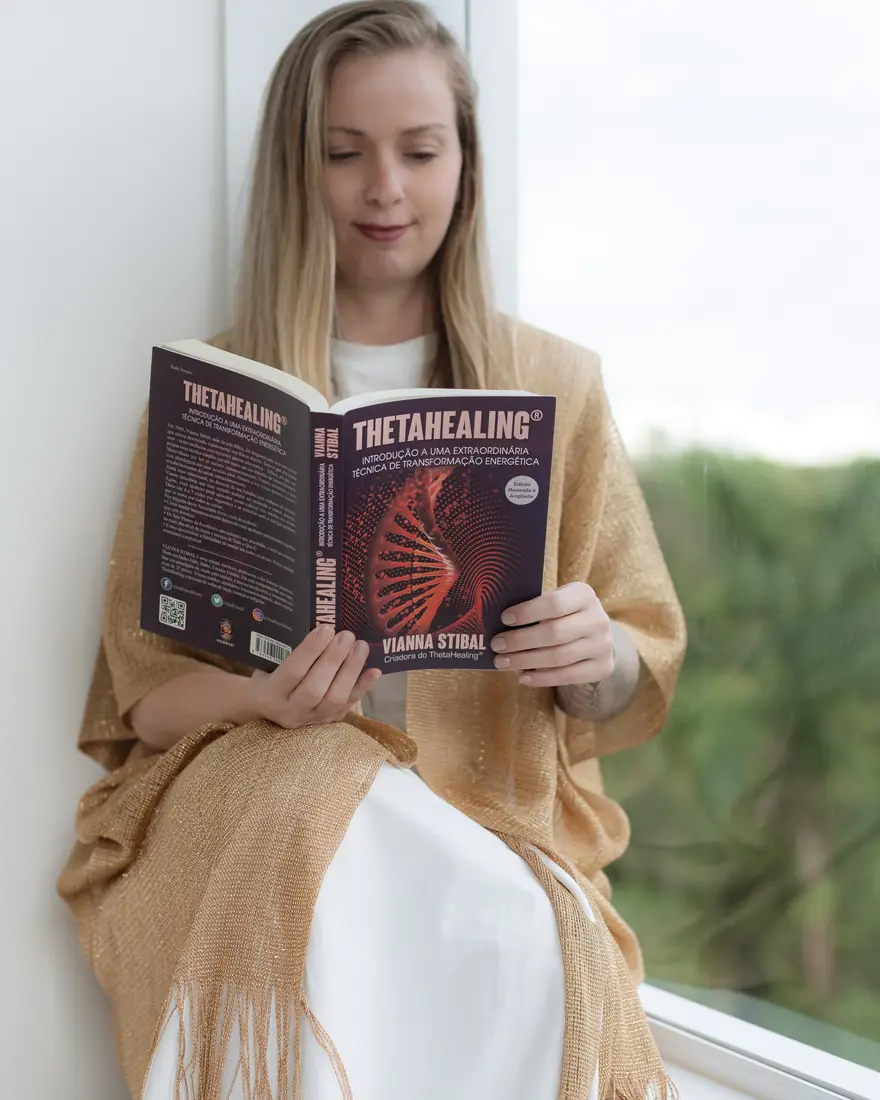 Lairany Souza sentada na janela lendo o livro ThetaHealing de Vianna Stibal